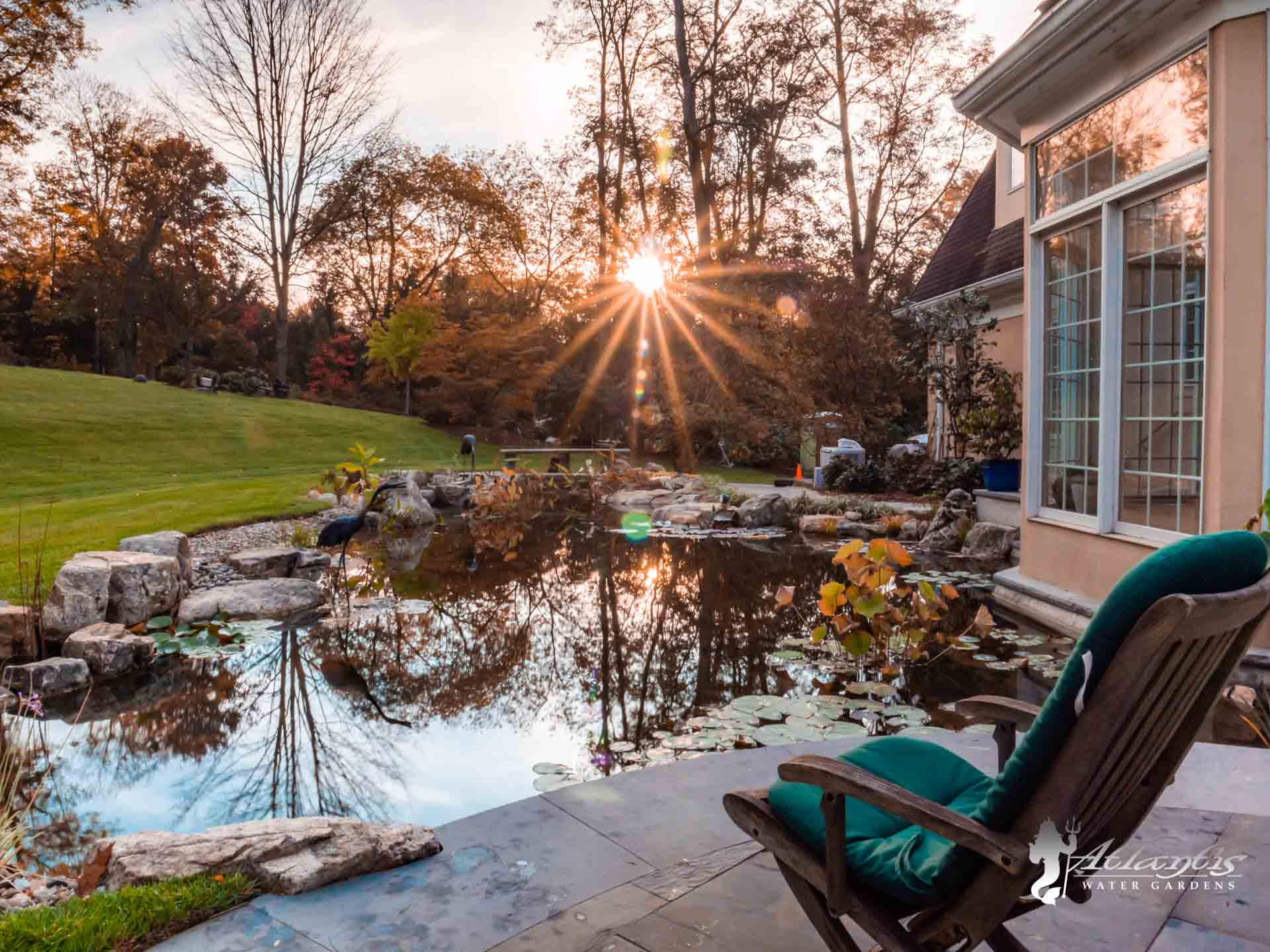 A serene backyard with a natural pond reflecting the sunset, surrounded by stonework and plants. There is a green cushioned chair on a stone patio next to a house with large windows